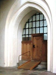 Indgangsparti til kirke med to trin op til døren og trærampe med håndliste.