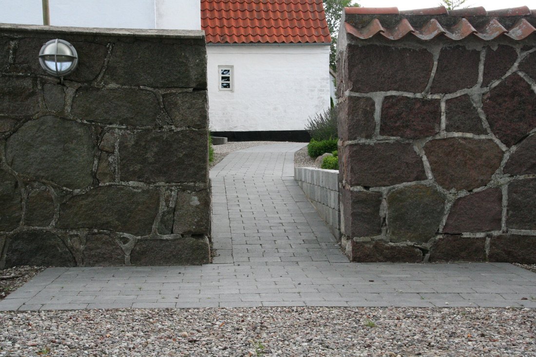 Sti mellem to halvmure fra parkeringsplads til bygning. Stien består af fliser, lagt i et rektangel foran murene samt på selve stien ind til bygningen.