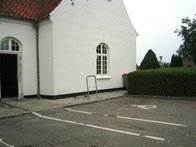 Parkeringsplads med kørestolsbruger malet på asfalten foran hvid kirke.
