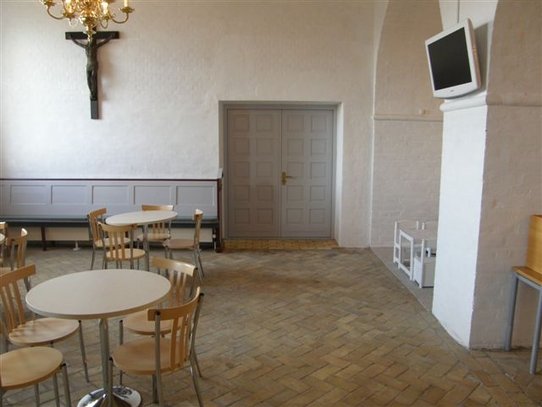 Kirkerum med rundborde, dobbelt indgangsdøre, samt fladskærm ophængt på væg.